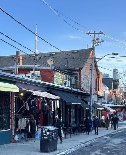 A vibrant street scene featuring various shops displaying clothing on hangers. The background includes a prominent tower and power lines against a clear blue sky. People are walking along the sidewalk, with some seated at outdoor tables. There is visible urban artwork on buildings.