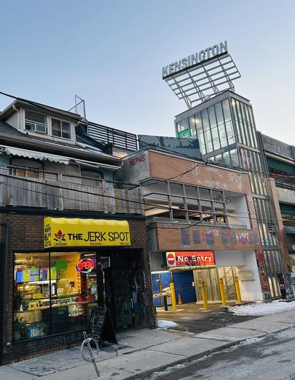 The image features a streetscape in Kensington with a bright yellow storefront for "The Jerk Spot," showcasing an open sign. The building above shows various signs of graffiti and a prominent Kensington sign