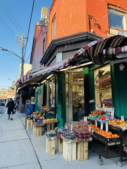 A colorful fruit market features various fruits displayed on wooden crates outside. The storefront has a green facade with striped awnings, while a person walks by on the sidewalk. The background shows a clear blue sky and urban surroundings.