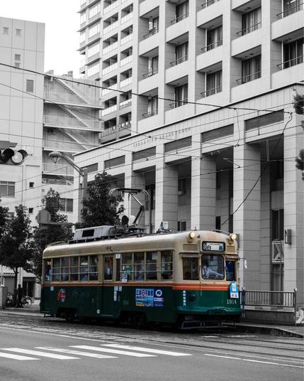 A vintage Japanese streetcar numbered 1914 in a selective color photograph. The streetcar features a cream-colored upper body and dark green lower section, standing out against a black and white urban backdrop. The tram is positioned in front of a modern high-rise apartment building with a distinctive zigzag fire escape. The streetcar has an overhead electric line and runs on traditional tracks embedded in the street. The scene combines classic public transport with contemporary Japanese urban architecture, creating a striking contrast between old and new.
