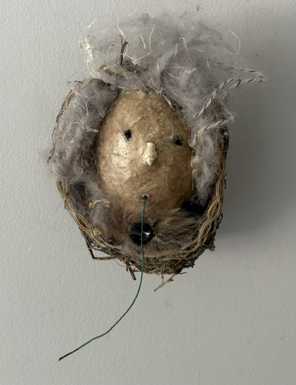 A paper maché mask with small black stones for eyes and a tiny shell for a nose has a piece of green string hanging from its mouth. It is mounted on a small birds nest, half the size of a robins nest - maybe a wren? The mask has a lot of fluffed up grey wool for hair and a bit of fur tucked in at the bottom.