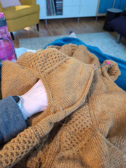 Seen from my point of view, a dark yellow cardigan being knit is draped on my lap. My left hand is in one of the pockets to show its depth. 