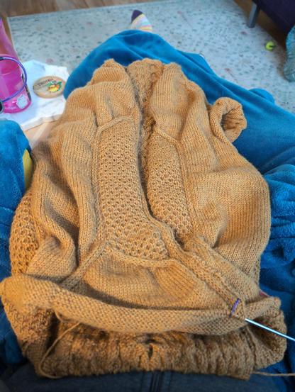 From my point of view, a full view of the body of a dark yellow cardigan being knit draped on my lap. It features cables and pockets on the front. The sleeves are still missing. 