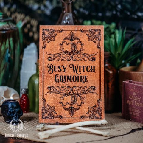 A brown journal with a leather texture is propped up among animal bones and burning black skull candle. The black text on the cover reads "Busy Witch Grimoire," surrounded by black Baroque embellishments. This is an exclusive product from Inked Goddess Creations.