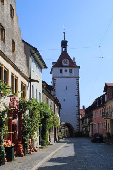 Blick entlang einer Straße mit Kopfsteinpflaster. Links und rechts Häuserzeilen, teils farbig gestrichene Häuser, teils berankt. Am Ende der Straße befindet sich ein weißer, quadratischer Stadtturm mit einem Rundbogen-Tor.
