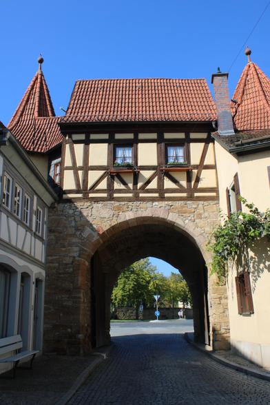Blick auf ein Stadttor mit Rundbogentor. Der untere Teil ist aus Naturstein, oben Fachwerk mit hellgelben Gefachen. Links und rechts sind Türmchen zu erkennen. 