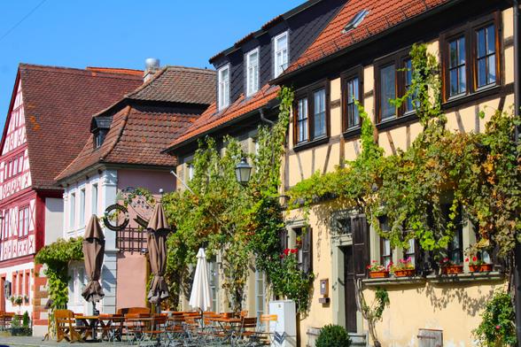 Blick auf eine Häuserzeile: rotes Fachwerk, Haus in hellblau-weiß, stark beranktes Gebäude mit geschlossenen Sonnenschirmen und leeren, roten Klappstühlen davor. Vorne ein hellgelbes Fachwerkhaus mit Wein berankt. Der Himmel ist blau, die Atmosphäre freundlich-sommerlich.