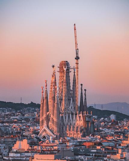 Eine Nahaufnahme der Sagrada Família in der Abendsonne. Die kunstvollen Türme der Basilika leuchten in warmem Orangeton, während sich ihre komplexen Details und Verzierungen abzeichnen. Ein hoher Baukran deutet darauf hin, dass die Kirche weiterhin im Bau ist. Die umliegende Stadtlandschaft mit ihren Gebäuden und Straßen breitet sich in alle Richtungen aus, während im Hintergrund Hügel und ein pastellfarbener Himmel zu sehen sind.