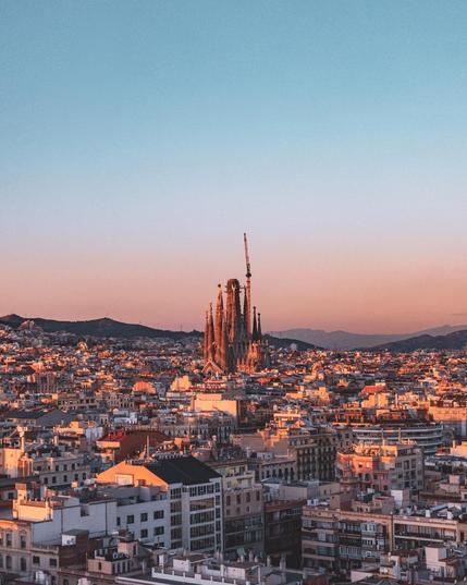 Das Bild zeigt eine weitläufige Stadtlandschaft in der Abenddämmerung, über der ein klarer Himmel mit einem sanften Farbverlauf von Blau zu Rosa liegt. In der Mitte ragt die berühmte Basilika Sagrada Família mit ihren kunstvollen Türmen und Baukränen in die Höhe. Die umliegenden Gebäude sind dicht gedrängt und reflektieren das warme, goldene Licht der untergehenden Sonne. Im Hintergrund sind sanfte Hügel zu erkennen, die die Stadt umgeben.