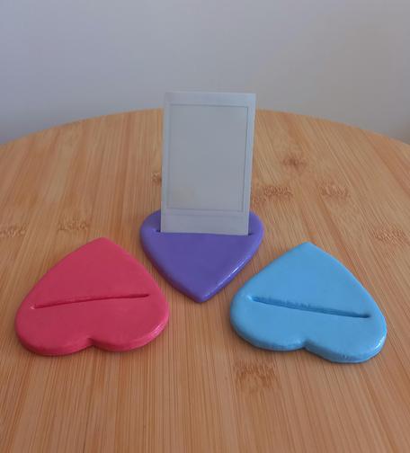 Foto a tres portafotos planos en forma de corazón sobre una mesilla de madera. El de la izquierda es rosa, el del medio es morado y en él hay una foto Polaroid blanca vertical, y el de la derecha es azul.