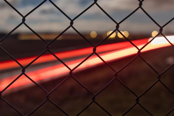 Description : Un grillage métallique en premier plan, net et figé, séparant l’observateur d’un monde en mouvement. Derrière, des traînées lumineuses rouges et blanches fendent l’obscurité, témoins silencieux d’une vitesse qui échappe. Des points dorés flottent au loin comme des étoiles perdues. Entre barrière et évasion, la lumière danse, emprisonnée sans l’être vraiment.  

Poésie : Les lueurs s’entrelacent derrière les mailles, la lumière file sans entrave, tandis que l’ombre demeure presque immobile. L’éphémère traverse le figé, comme une promesse d’ailleurs. Parfois, c’est le regard qui choisit : prison ou passage, enfermement ou évasion. 