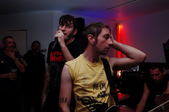 Un concert de punk, le chanteur tient le micro à sa bouche, le guitariste porte un t-shirt jaune et semble tenir la pose. En fond des néons bleus et rouge.