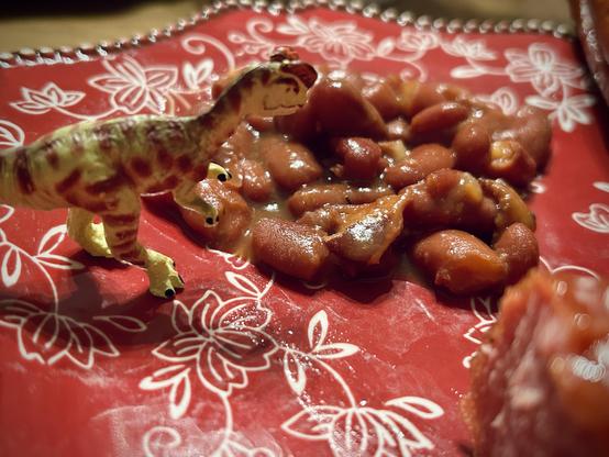 A green Dilophosaurus with brown spots stands on a red plate decorated with white floral patterns. They stand before a pile of brown baked beans. A kielbasa is in the foreground and partially blurred.