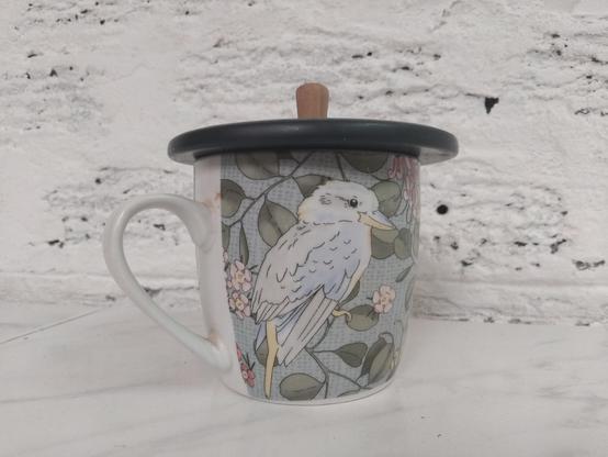 A cup or small mug with a kookaburra design on it and a grey lid, of 15% greater diameter than the cup, with a small wooden knob for a handle, sitting on top of the cup.