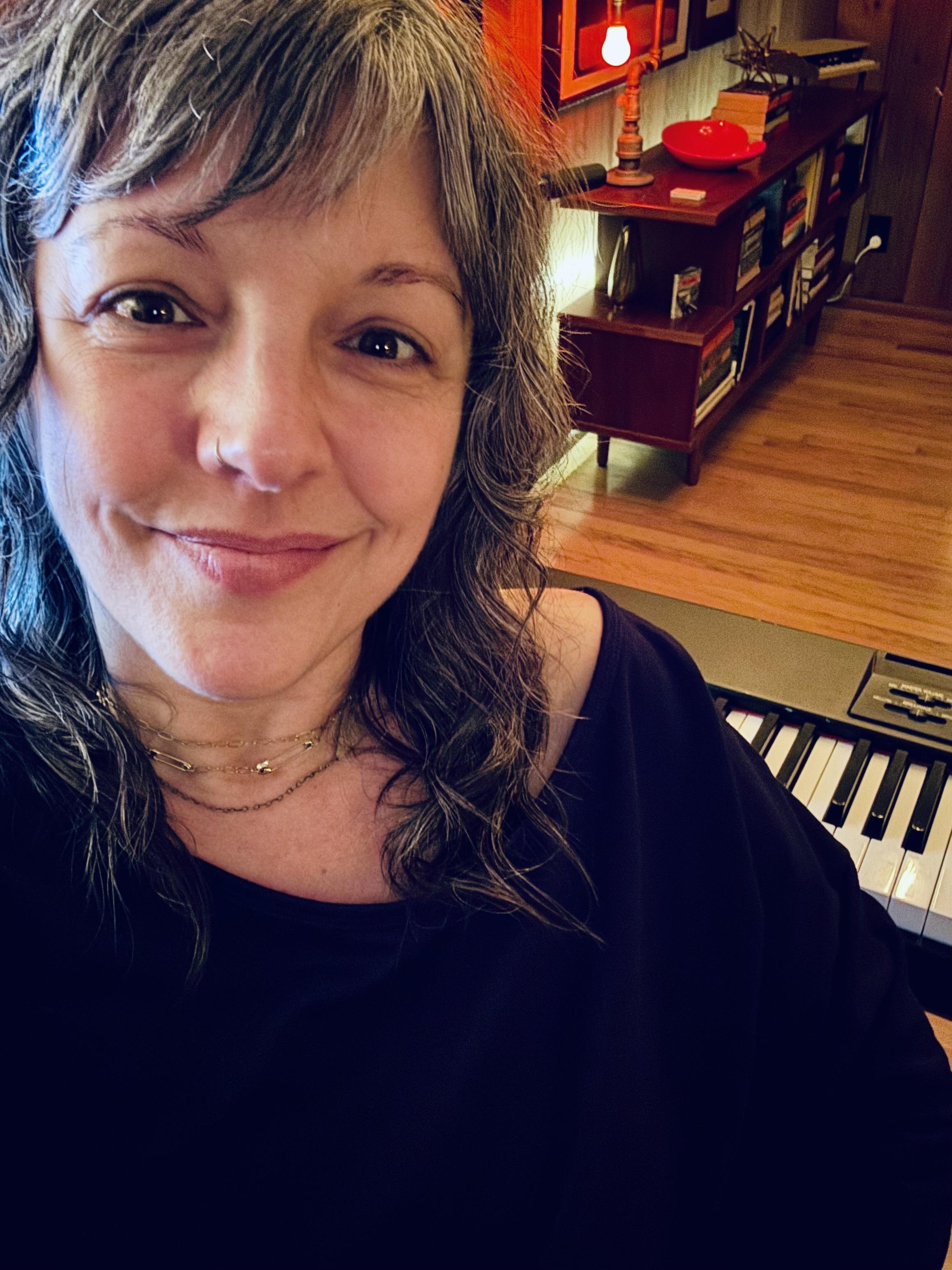 This is a selfie style photograph of Shannon sitting in her studio. There is a keyboard visible behind her shoulder and beyond that there is a low bookcase with books and other colorful tchotchkes on it.