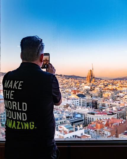 Ein Mann mit kurzen, dunklen Haaren steht mit dem Rücken zur Kamera vor einem großen Fenster mit Blick auf eine Stadtlandschaft. Er trägt ein schwarzes T-Shirt mit der Aufschrift „MAKE THE WORLD SOUND AMAZING.“ in weißen und grünen Buchstaben. Er hält ein Smartphone in beiden Händen und macht ein Foto der Stadt. Im Hintergrund erstreckt sich eine dichte Bebauung mit vielen Gebäuden, und im Zentrum ragt die berühmte Sagrada Família in Barcelona empor. Der Himmel zeigt einen sanften Farbverlauf von Blau zu einem warmen Orangeton, was auf Sonnuntergang hindeutet. Das Fenster hat leichte Verschmutzungen oder Kratzer.