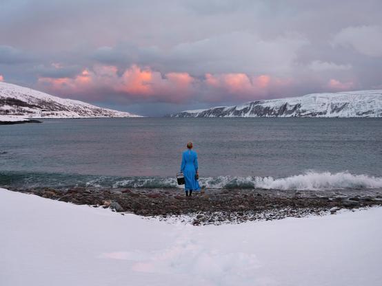Arktische Fjordlandschaft mit dramatischen Wolken, am Strand steht eine Frau in einem blauen Kleid, die man von hinten sieht.