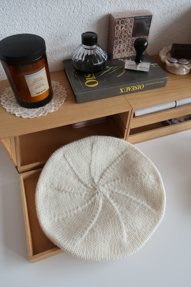 Un béret en laine couleur crème vue d’en-dessus, posé sur une petite commode. En arrière plan, se trouvent un livre sur les oiseaux, une bougie et des pins dans une coupelle.
