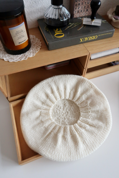 Un béret en laine couleur crème vue d’en-dessous. On voit l’intérieur du béret, avec la bordure élastique. Eposé sur une petite commode. En arrière plan, se trouvent un livre sur les oiseaux, une bougie et des pins dans une coupelle.