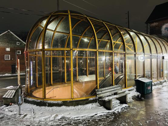 A modern, glass-enclosed transit shelter illuminated at night, with an escalator visible inside. Snow covers the ground, and a bench and a green trash bin are placed outside the structure.