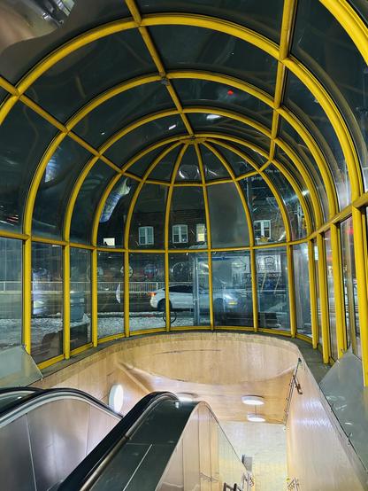 An escalator descends into a transit station enclosed by a glass and yellow framework. The scene is illuminated with artificial lighting, while snow is visible outside along with vehicles passing by.