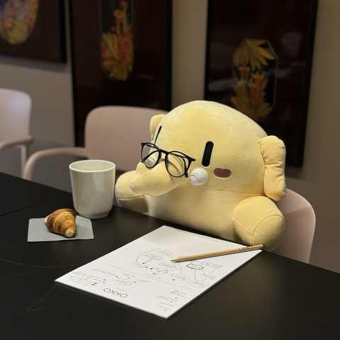 Mastodon’s plushie sitting at a table with folded glasses on his trunk, coffee mug and a small croissant from one side and a notebook with a pencil from another.