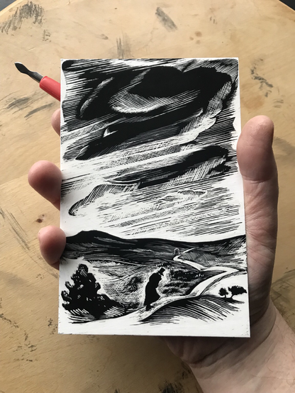 Photo of a hand over a wooden surface holding a sharp tool with a red handle and a small card with a black and white design upon it. The design shows a landscape with trees and hills, a brooding sky and a path stretching off into the distance to a small structure. A lone figure in a large coat and top hat strides towards it