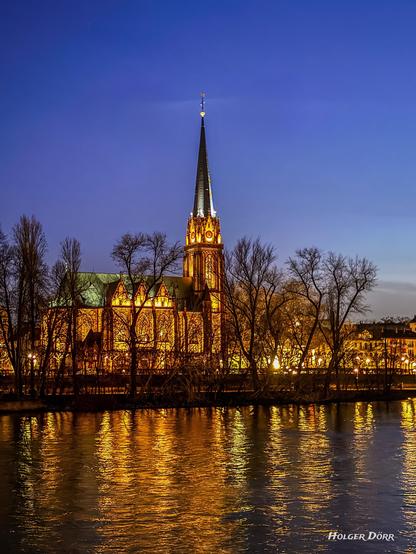 Eine stimmungsvolle Abendaufnahme der Dreikönigskirche in Frankfurt am Main.
Die Kirche leuchtet golden im künstlichen Licht, während sich ihre Spiegelung im sanft bewegten Wasser des Mains widerspiegelt. Der Himmel ist tiefblau und kahle Bäume im Vordergrund rahmen die Szenerie ein. Die Aufnahme fängt die Magie der Blauen Stunde ein und verleiht der Szene eine märchenhafte Atmosphäre.