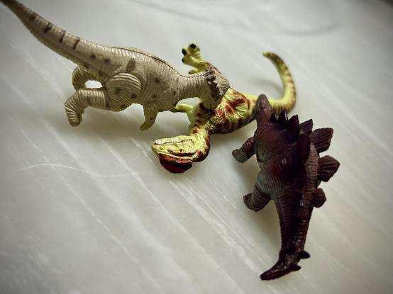 A green Dilophosaurus with brown spots lies on a white marble surface and is eaten by a tan Pachycephalosaurus and red Stegosaurus.