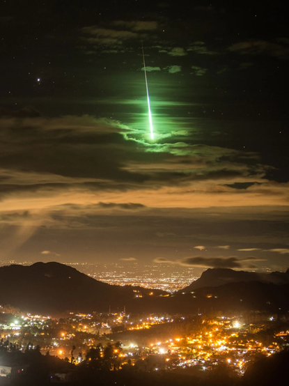 Bright green streak plunging through clouds above a sea of city lights.