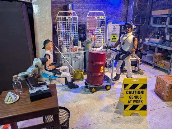 Close shot of the mad science set.  Minion #1 sits in a chair at left, mop and bucked at hand. Minion Number Nine pushes a dolly with a large red barrel with a radioactive symbol on it. I floor tent sign reads, "CAUTION! GENIUS AT WORK" In the background, lab benches with various chemistry and science items on them, and a large white locker, also marked with a radiation symbol.