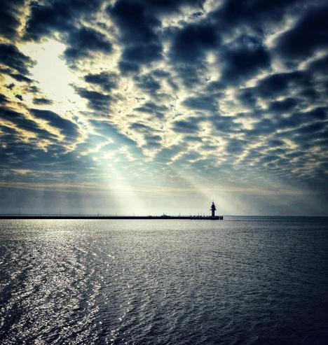 This photo captures a stunning seascape with a dramatic sky filled with textured clouds. The sun's rays break through the gaps in the clouds, casting beams of light onto the water below. The ocean surface is calm, with gentle ripples reflecting the sunlight. In the distance, a long pier extends across the horizon, leading to a solitary lighthouse standing at the edge. The contrast between the dark clouds and the bright sunbeams creates a striking and atmospheric scene.