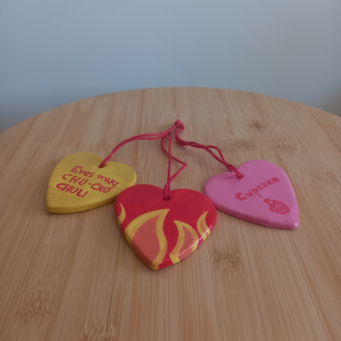 Foto en la que aparecen 3 colgantes en forma de corazón con un hilo rojo de yute anudado cada uno sobre una mesilla de madera. Se ve su parte delantera. El de la izquierda es amarillo y tiene escrito en rojo "Eres muy CHU-CHU-CHULI", el del medio es rojo con llamas en su parte inferior y el de la derecha es rosa con la palabra "Cupcake (pastelito)" escrita en rojo y un pastelito con un sombrero de guinda dibujado en rojo.