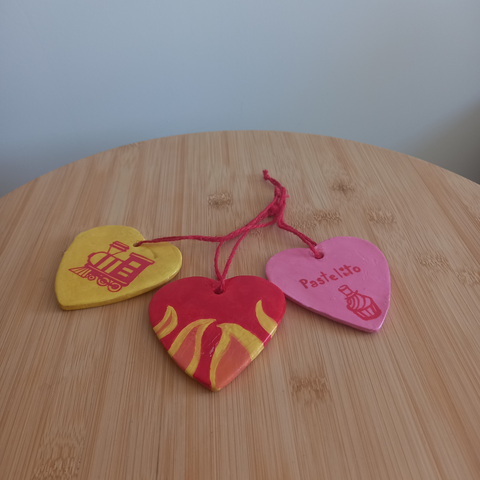 Foto en la que aparecen 3 colgantes en forma de corazón con un hilo rojo de yute anudado cada uno sobre una mesilla de madera. Se ve su parte trasera. El de la izquierda es amarillo y tiene dibujado en rojo un tren (una locomotora), el del medio es rojo con llamas en su parte inferior y el de la derecha es rosa con la palabra "Pastelito" escrita en rojo y un pastelito con un sombrero de guinda dibujado en rojo.