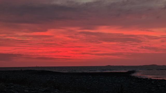 Historical Documentation in the form of a photograph of a rising over the ocean in Winthrop, MA.  The sky is filled with vibrant red and orange hues, with the sun rising on the horizon and casting a warm glow over the water. The clouds are scattered across the sky, creating a dramatic contrast with the cloud covered sky. The water is calm and reflects the colors of the sky. In the foreground, there is a rocky shoreline with small rocks and pebbles scattered across it. On the right side of the image, there are a few small islands visible in the distance. The overall mood of the photograph is peaceful and serene.