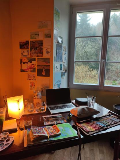 Desk with art supplies and a laptop and colorful cards and posters on the wall. The desk is facing a window looking out on the forest. There's a little triangular lamp on the desk with a yellowish light giving the wall an orange yellowy glow