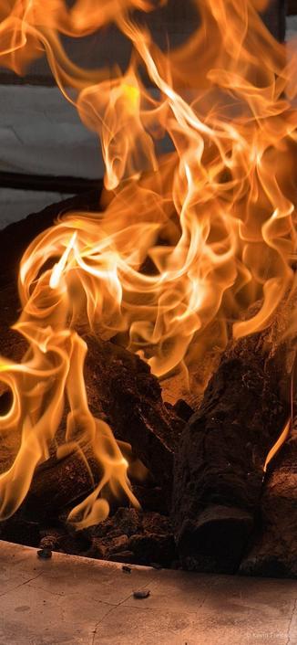 A close-up image of flames dancing over dark, charred wood logs, showcasing a vivid display of orange and yellow hues. The background is blurred, emphasizing the fiery texture and movement of the fire.