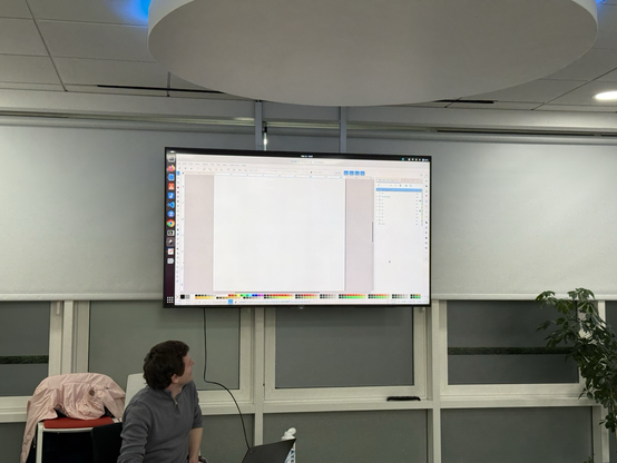 A person is sitting in front of a large screen displaying a digital drawing application. The workspace is mostly blank, with color palettes visible at the bottom. The setting appears to be a modern office environment.