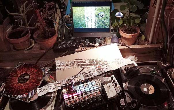 Photo de platines vinyles accompagnées de contrôleurs midi, d'une trace de pneu sur une feuille, et la reproduction de cette trace sur un écran d'ordinateur. ay 2em plan de cette trace, on voit un pneu, dressé verticalement dans un espace herbé. Posée sur les platines on voit aussi une bande de papier avec écrite une playliste de noms codés, et de Bpms... Il y a des plantes grasses autour de l'écran d'ordi. Autour il y a pas mal de câbles. Un pochoir de chaton en train de sauter sur un stiker posé sur un agenda.