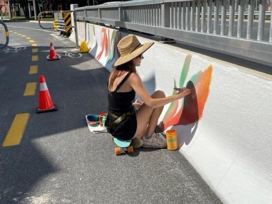 A woman is sitting on a small skateboard while painting a colorful mural on a concrete barrier. She is wearing a straw hat and a black outfit. There are traffic cones and a bike lane visible in the background.