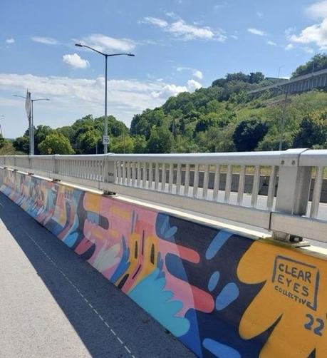 A colorful mural along a concrete barrier beside a road, featuring abstract shapes and patterns. The artwork is attributed to "Clear Eyes Collective" and is dated "22." In the background, there are green hills and blue skies with scattered clouds.