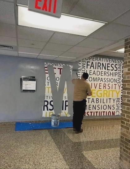 photo of a man using a paint roller to roll grey paint over a wall in a hallway, obliterating words like "Fairness" "Compassion" "Diversity" "Integrity" "Respect" etc