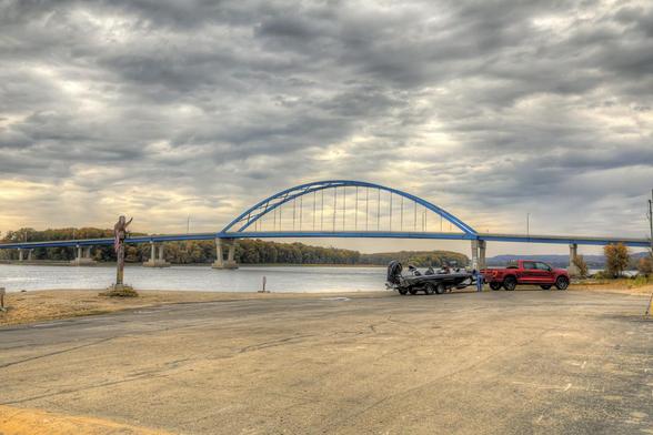 A red truck with a boat just removed from the water, on a trailer, sits parked near the Mississippi River, under a cloudy sky. 