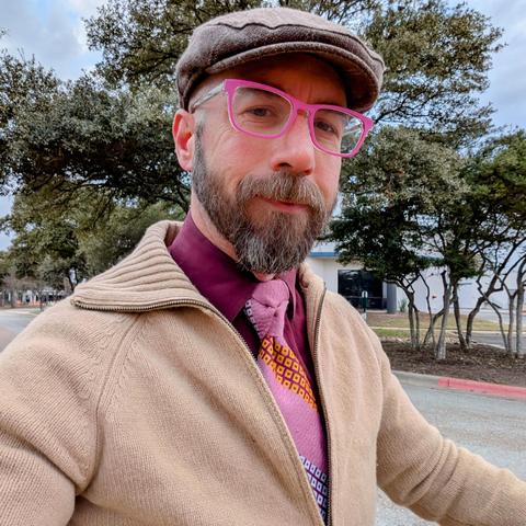 A photo of me, a 46 year old white man with a shaved head, a short cropped brown & grey beard, 

photographed from the chest up

In front of some live oak trees in an office park,

wearing

rectangular pink acetate glasses, a pink with diagonal stripes of white and yellow squares edged in maroon in an Eldredge Knot on maroon dress shirt under a camel cardigan with a light brown flat cap