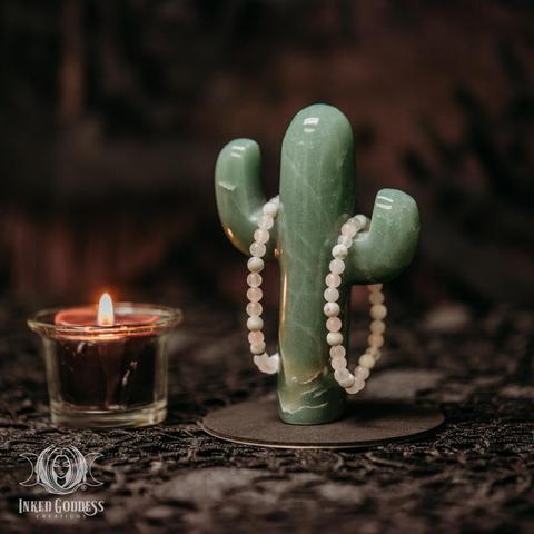 Two examples of the exclusive Love Gemstone Bracelet hang from a carved green aventurine saguaro cactus with a burning votive candle for scale. The bracelets are elastic and have no clasp. They consist of small round spherical beads of white howlite, white jade, and rose quartz. This is an exclusive product from Inked Goddess Creations.