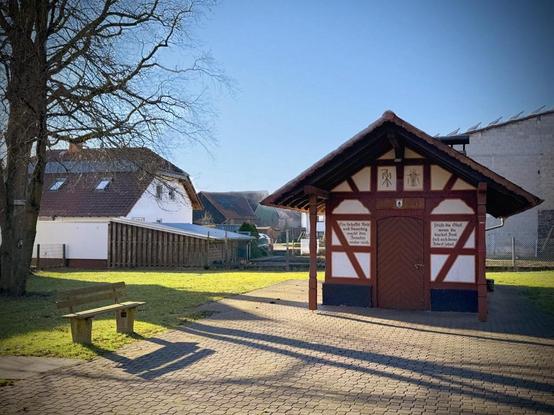 Das erste Foto zeigt das Backhaus in Münster bei Laubach in einer leicht versetzten Frontalansicht, wodurch auch das direkte Umfeld sichtbar ist.
Der Vorplatz des Backhauses ist mit Pflastersteinen belegt und geht in eine Grünfläche über. Eine Holzbank mit Betonfüßen steht links im Schatten eines großen Laubbaums, dessen Äste in den Himmel ragen. Im Hintergrund sind Wohnhäuser mit roten Dächern und moderne landwirtschaftliche Gebäude zu sehen. Der Himmel ist klar und blau, das Sonnenlicht wirft lange Schatten, was auf einen Wintertag oder frühen Frühling hindeutet. Die Szenerie vermittelt eine ruhige, dörfliche Atmosphäre.