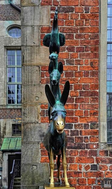 Ein Esel aus Metall steht vor einer Hauswand. An seiner Schnauze und seinen Beinen ist das Grün golden. Auf ihm steht ein Hund, auf diesem eine Katze, auf dieser ein Hahn.