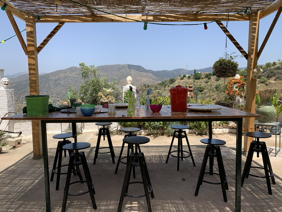 A la sombra de una pérgola de madera en exterior, una mesa grande con el material necesario para realizar piezas de barro. El paisaje montañoso de los montes de málaga a lo lejos está bañado por el sol de verano.