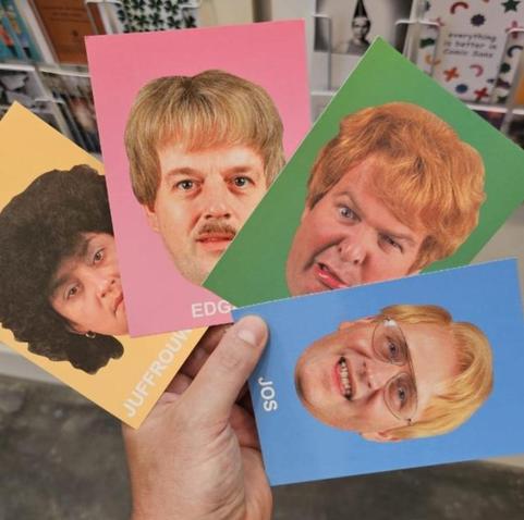 Een hand houdt vier ansichtkaarten vast met portretten van personages uit de Nederlandse comedyserie Debiteuren Crediteuren van Jiskefet. Op de kaarten staan de typetjes Edgar, Jos, Juffrouw Jannie en Storm, elk met een kenmerkend kapsel en gezichtsuitdrukking. De achtergronden van de kaarten zijn felgekleurd: roze, blauw, groen en geel. Op de achtergrond is een rek met andere kaarten en boeken zichtbaar.