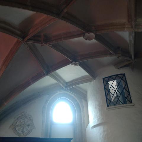 In der Mitte strömt helles Licht durch ein Fenster. Oben ist ein Kreutzrippengewölbe. Das Fenster rechts daneben ist vergittert, am Gitter ist ein Herz.